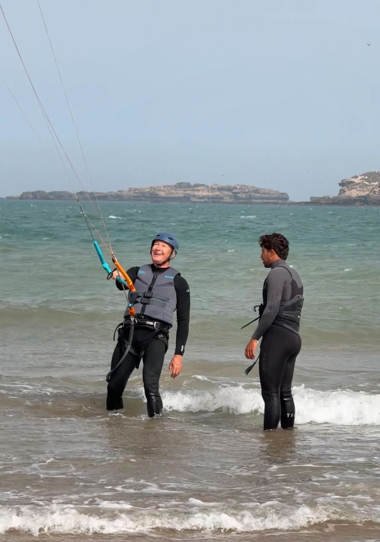Cours de kitesurf à Diabat, Essaouira — élève casqué apprenant à piloter l'aile dans l'eau avec son moniteur, îles de Mogador en arrière-plan, Windy Essaouira