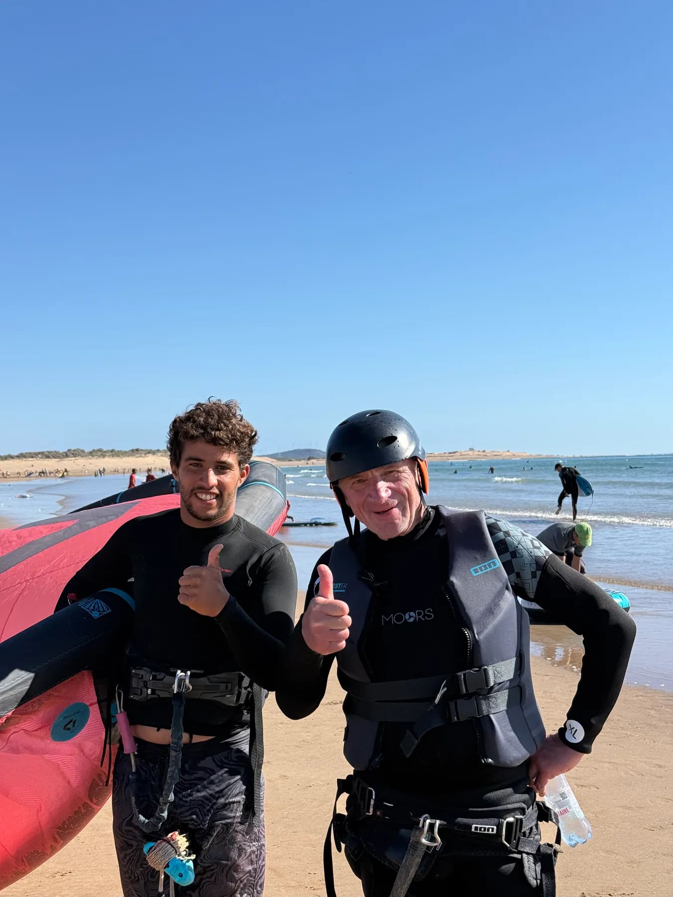Cours de kitesurf à Diabat, Essaouira — élève casqué et son moniteur pouce levé sur la plage après la session, aile posée sur le sable, Windy Essaouira