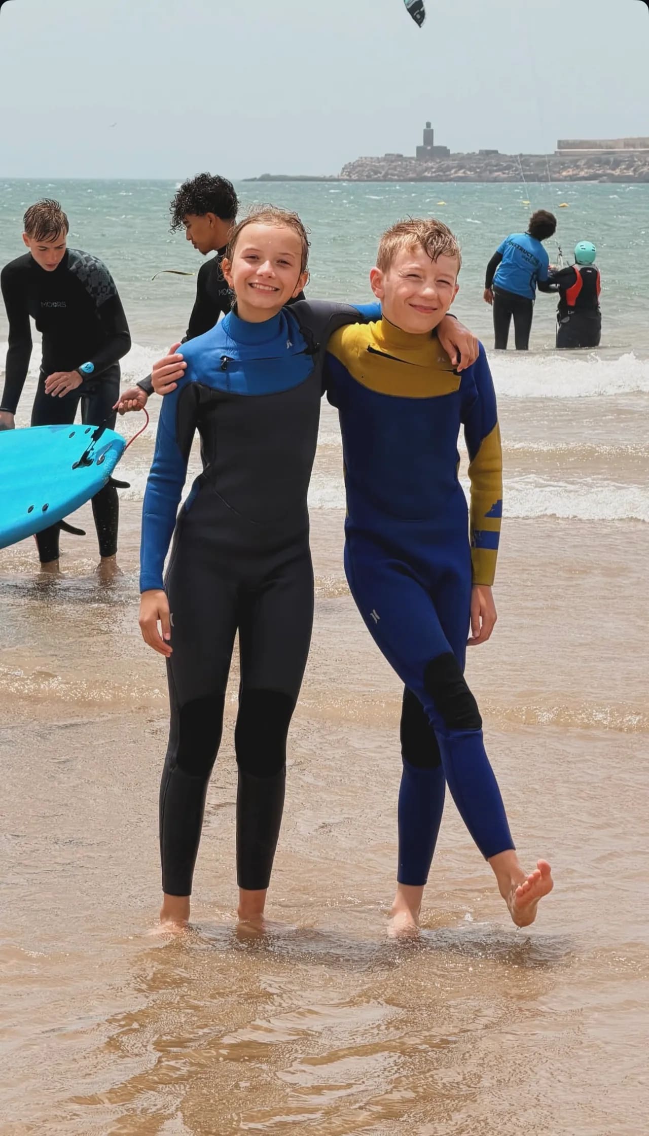 Two smiling kids in wetsuits on Essaouira beach after their surf lesson, other students and instructor behind — Windy Essaouira