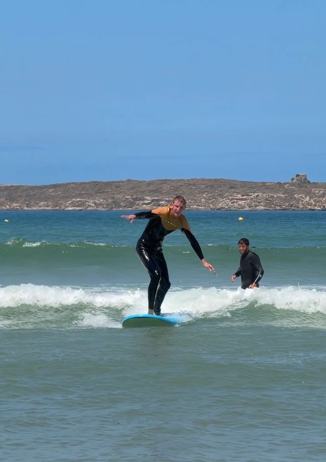 Élève debout sur sa planche lors d'un cours de surf à Essaouira, moniteur souriant derrière dans les vagues — école Windy Essaouira